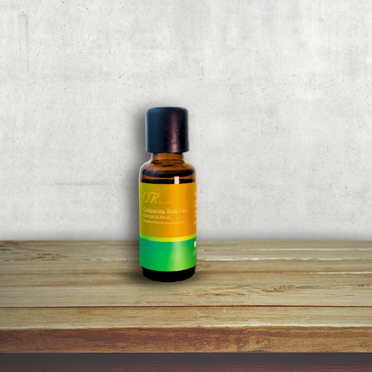 A small amber glass bottle of hair oil with a black cap, featuring a yellow and green label, standing on a wooden surface against a textured gray wall.