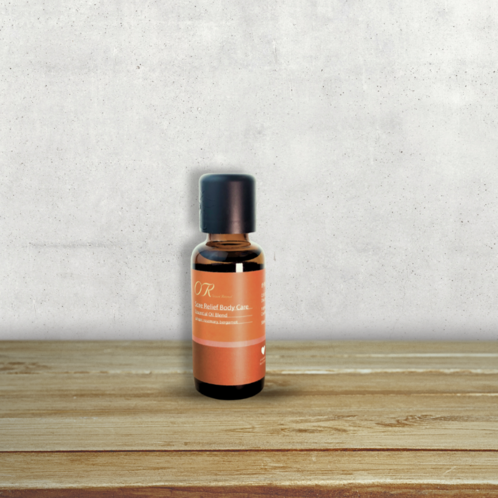 A small amber glass bottle with a black cap and an orange label reading 'OR' and 'Rose Herbal Balms', standing on a wooden surface against a textured light gray wall.