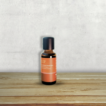 A small amber glass bottle with a black cap and an orange label reading 'OR' and 'Rose Herbal Balms', standing on a wooden surface against a textured light gray wall.