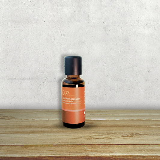 A small amber glass bottle with a black cap and an orange label reading 'OR' and 'Rose Herbal Balms', standing on a wooden surface against a textured light gray wall.
