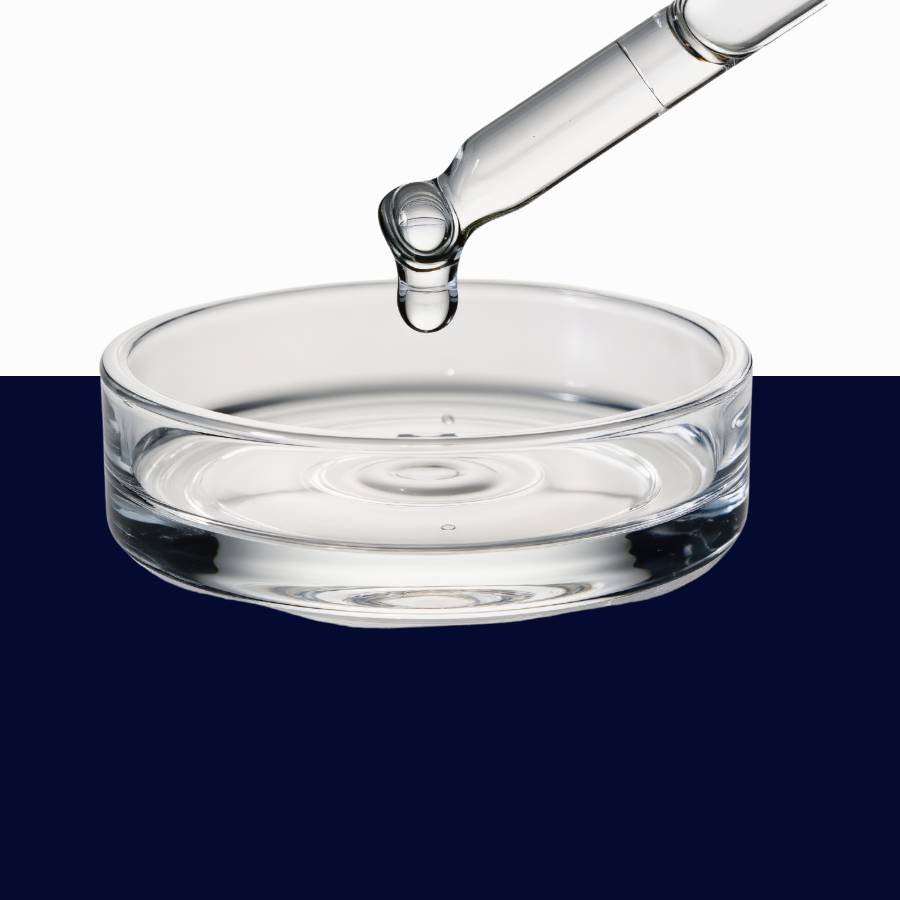 A clear glass pipette dripping transparent liquid into a shallow round glass dish, shown against a split white and dark navy background.