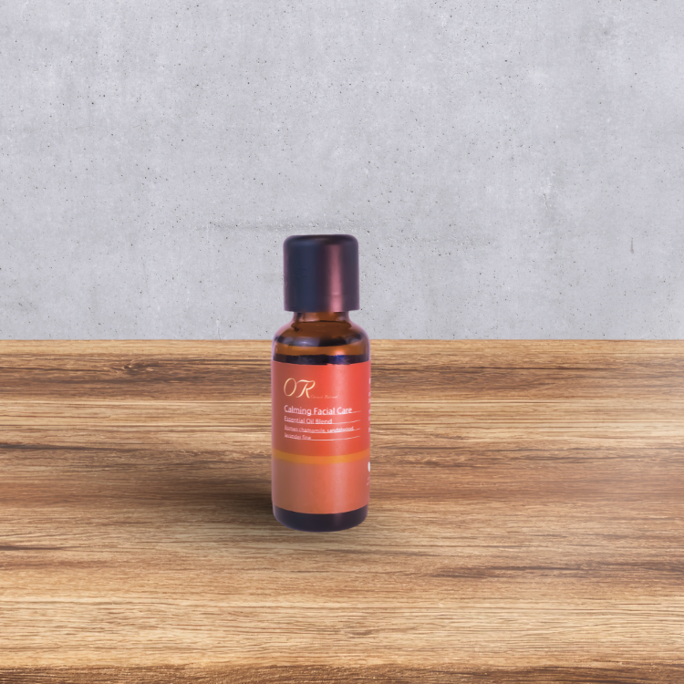A small amber glass bottle with a dark cap and a terracotta-colored label reading 'OR Coming Facial Care' stands centered on a wooden surface against a gray concrete wall.