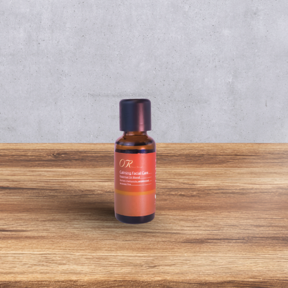A small amber glass bottle with a dark cap and a terracotta-colored label reading 'OR Coming Facial Care' stands centered on a wooden surface against a gray concrete wall.