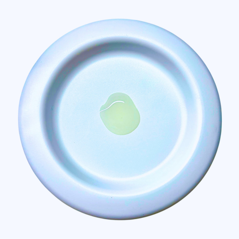 A circular white ceramic dish or saucer with a small dollop of pale yellow-green liquid in the center, set against a plain white background.